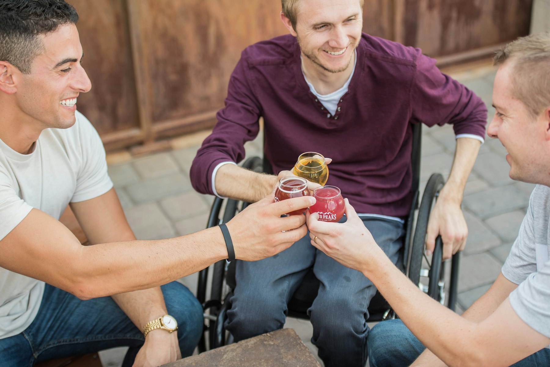 Three friends laughing together, one using a wheelchair, sharing a snack outdoors